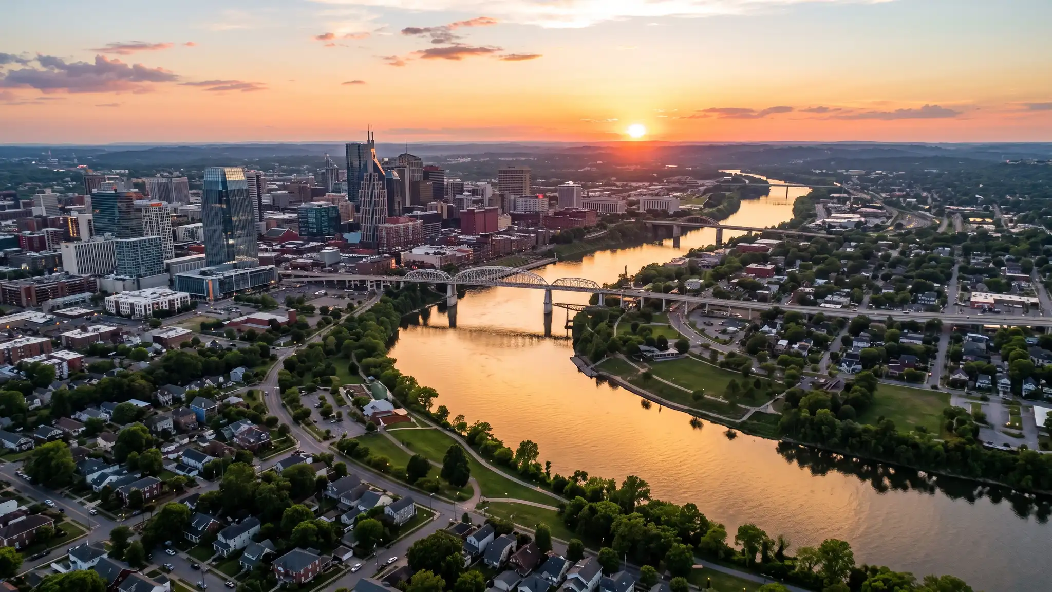 Aerial view of the Cumberland River winding past the Nashville skyline and residential neighborhoods at sunset with golden light on the water